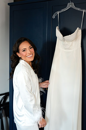 Bride in pajamas stands next to hanging wedding dress, smiling