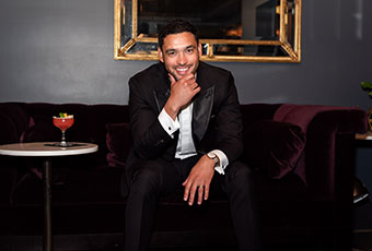 The groom, sitting and smiling at the camera, a cocktail is next to him