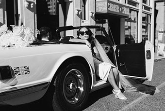 Bride sits in convertible with door open, legs out, sunglasses on