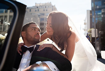 Bride hugs groom from behind. Groom is in drivers seat of car
