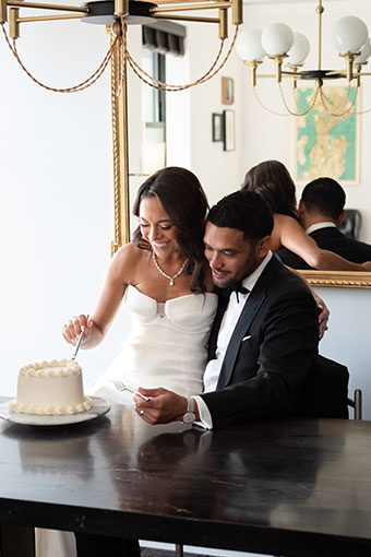 Bride sits on groom's lap as they both take use forks to take bite out of Just Married cake
