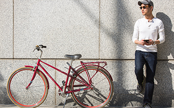 Man wearing a hat and sunglasses stands with a leg against a wall, next to a red bike